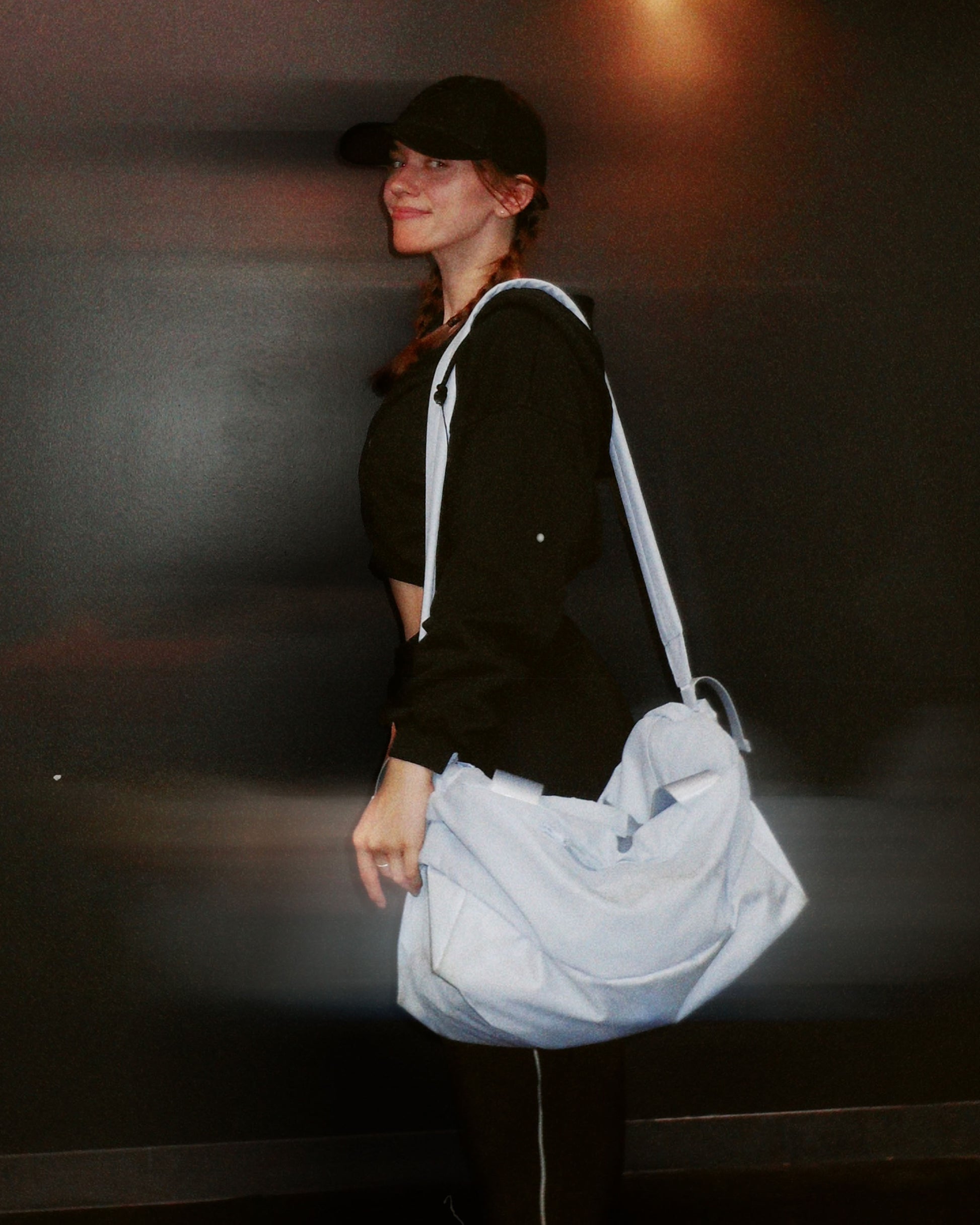 person wearing a black cap and black top, holding a light blue upper gym bag against a dark background