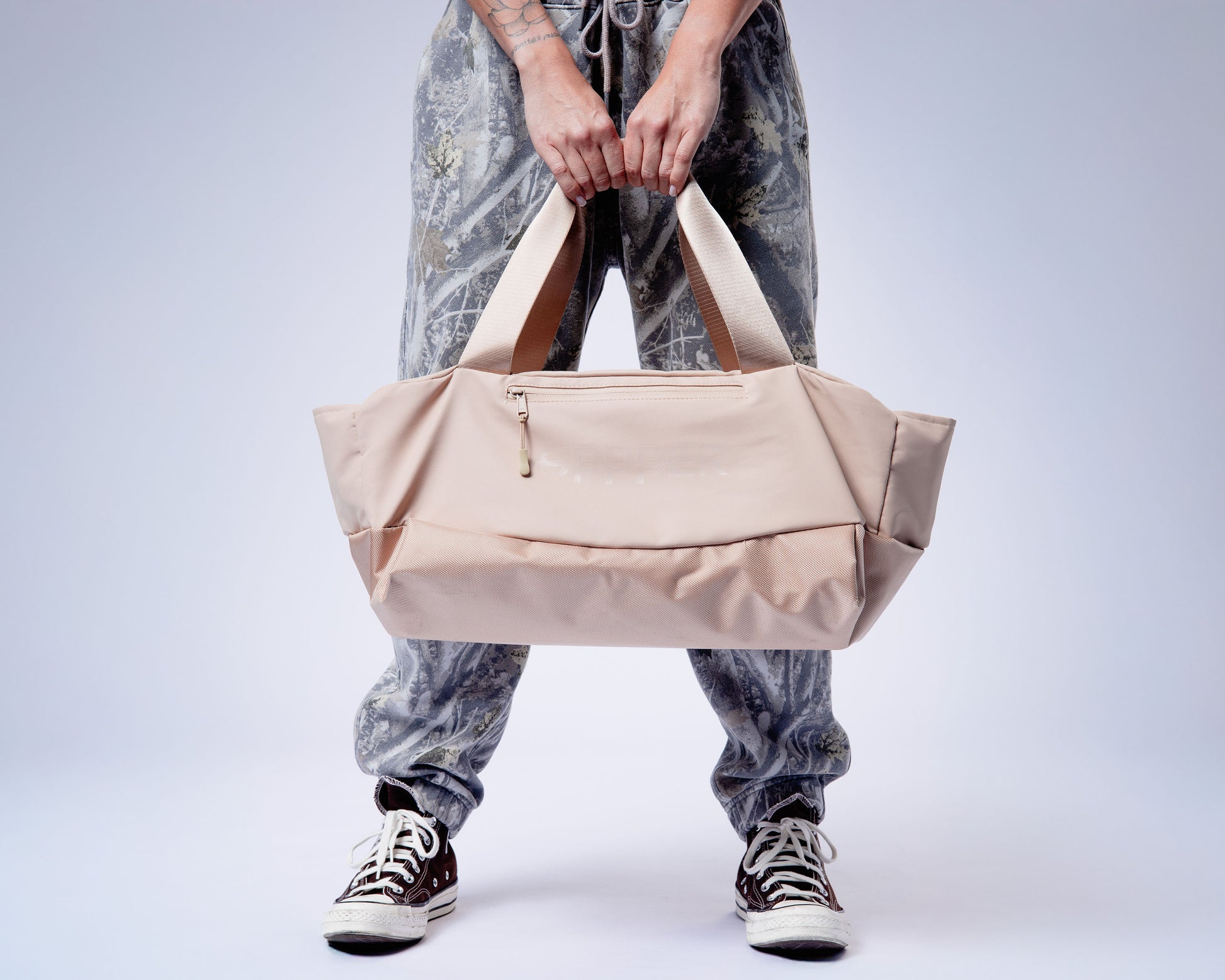 person holding a beige duffel uppper gym bag against a plain background
