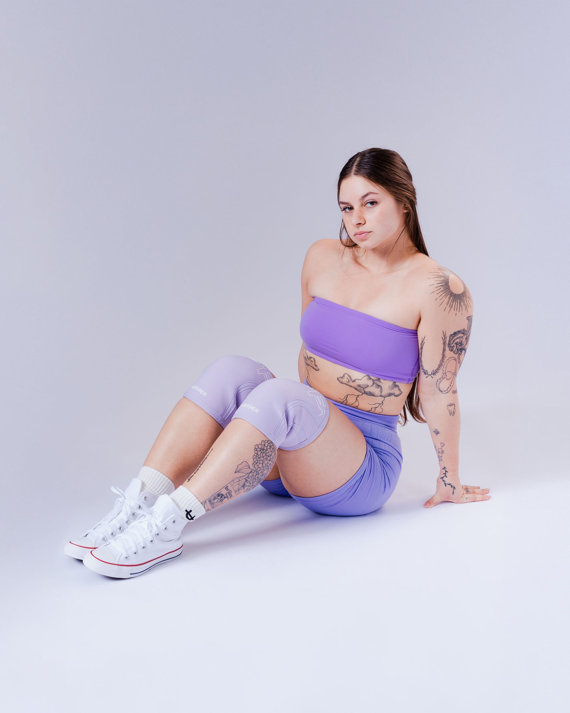 woman posing with lavender uppper knee sleeves wearing a purple outfit with white sneakers on a light gray background