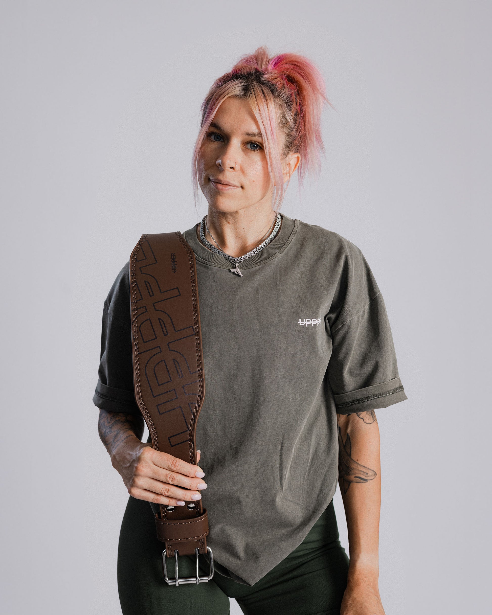 model wearing a gray shirt with uppper brand logo, holding uppper lifting belt in umber color against a plain background
