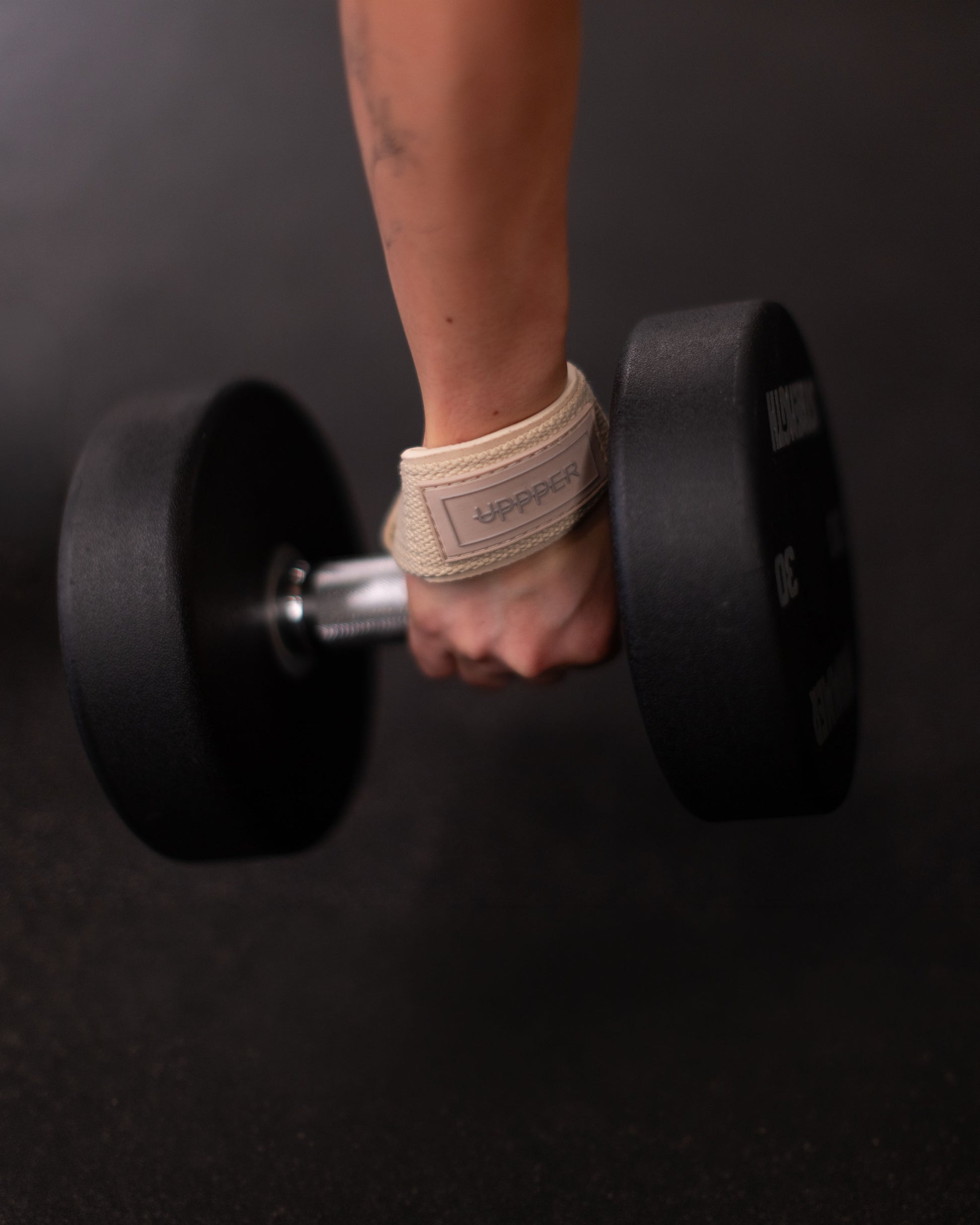 hand holding a black dumbbell with upper lifting straps in neutral color on dark background